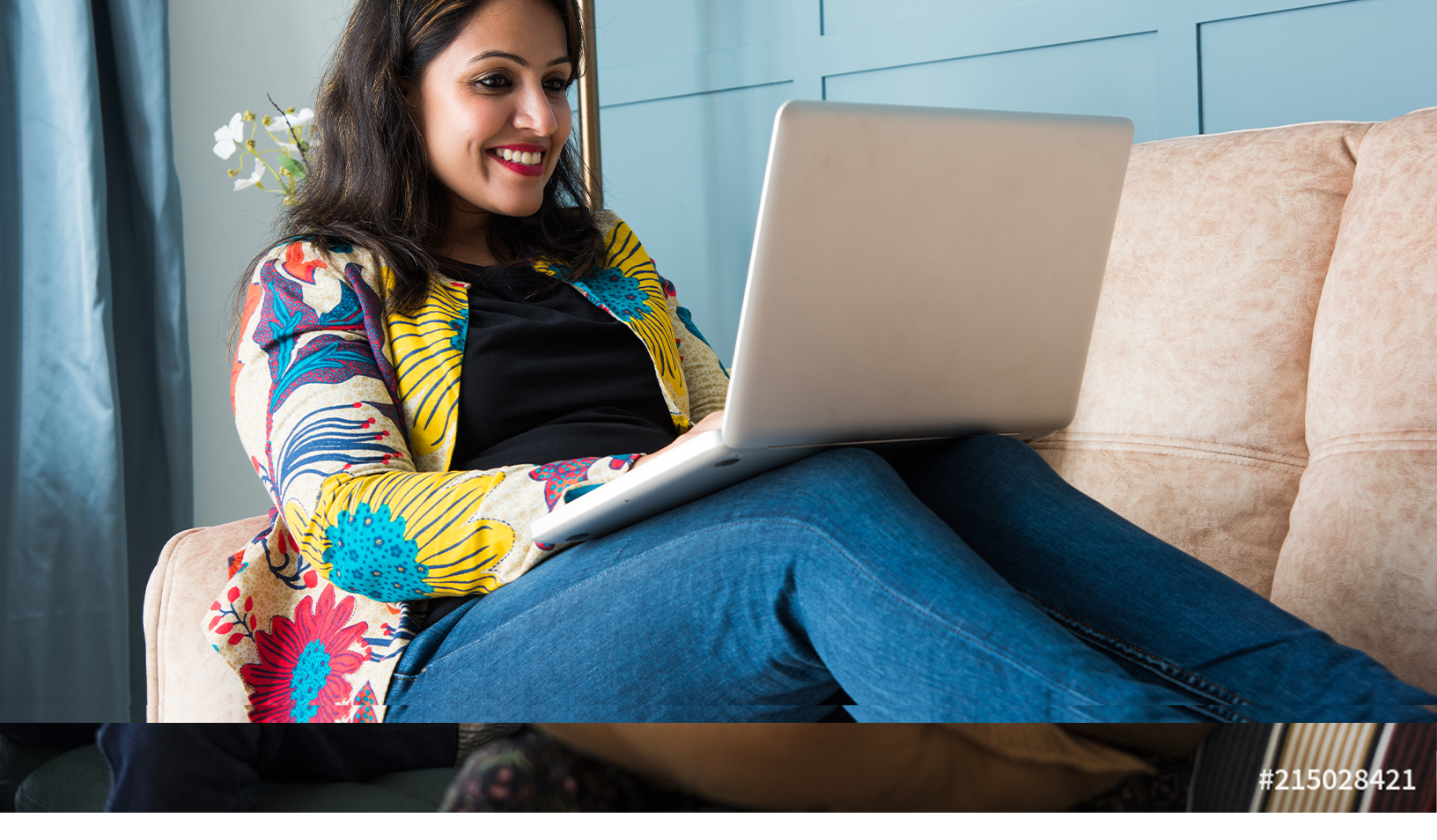 A woman sits comfortably on a couch, smiling while using a laptop. She is wearing a colorful jacket and blue jeans. A vase with white flowers is visible in the background.