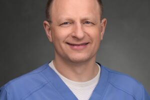 A middle-aged man with short hair is wearing light blue medical scrubs and a white undershirt, posing against a plain, dark background.