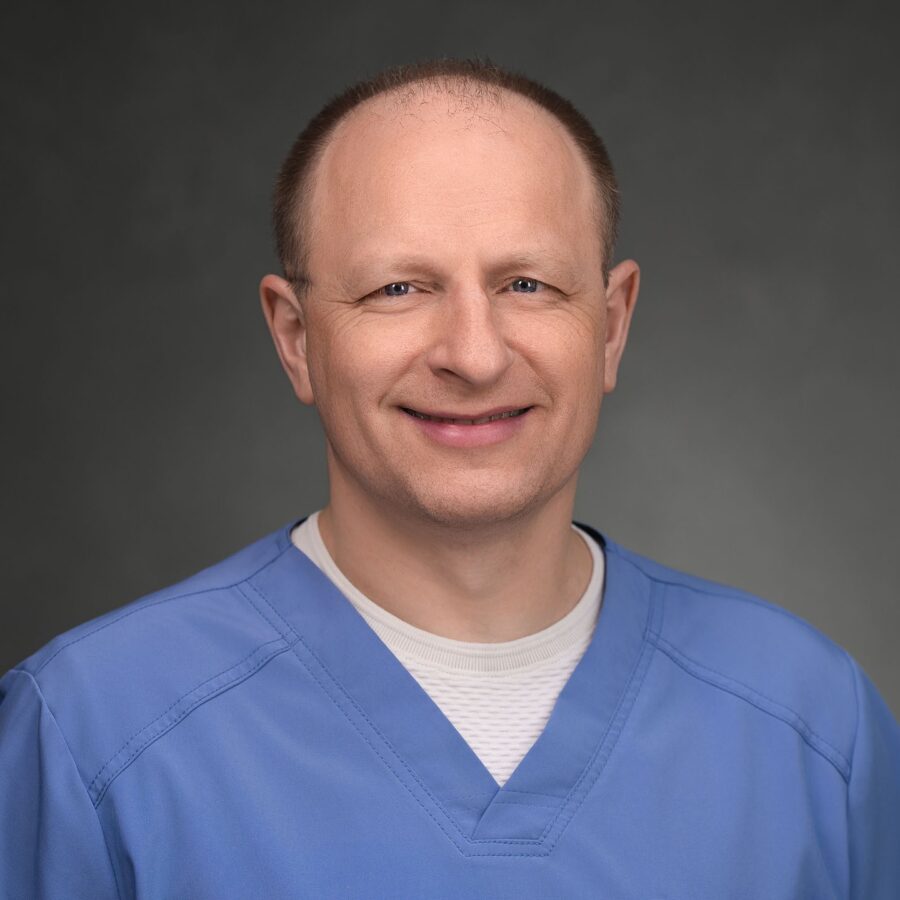 A middle-aged man with short hair is wearing light blue medical scrubs and a white undershirt, posing against a plain, dark background.