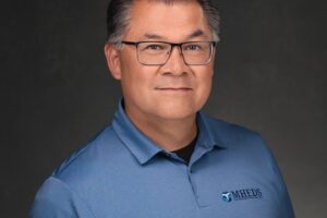 A man with short gray hair and glasses wears a blue polo shirt with an MHEDS logo, posing against a dark background.