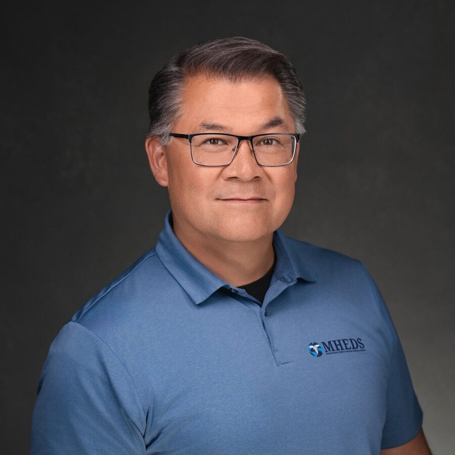 A man with short gray hair and glasses wears a blue polo shirt with an MHEDS logo, posing against a dark background.