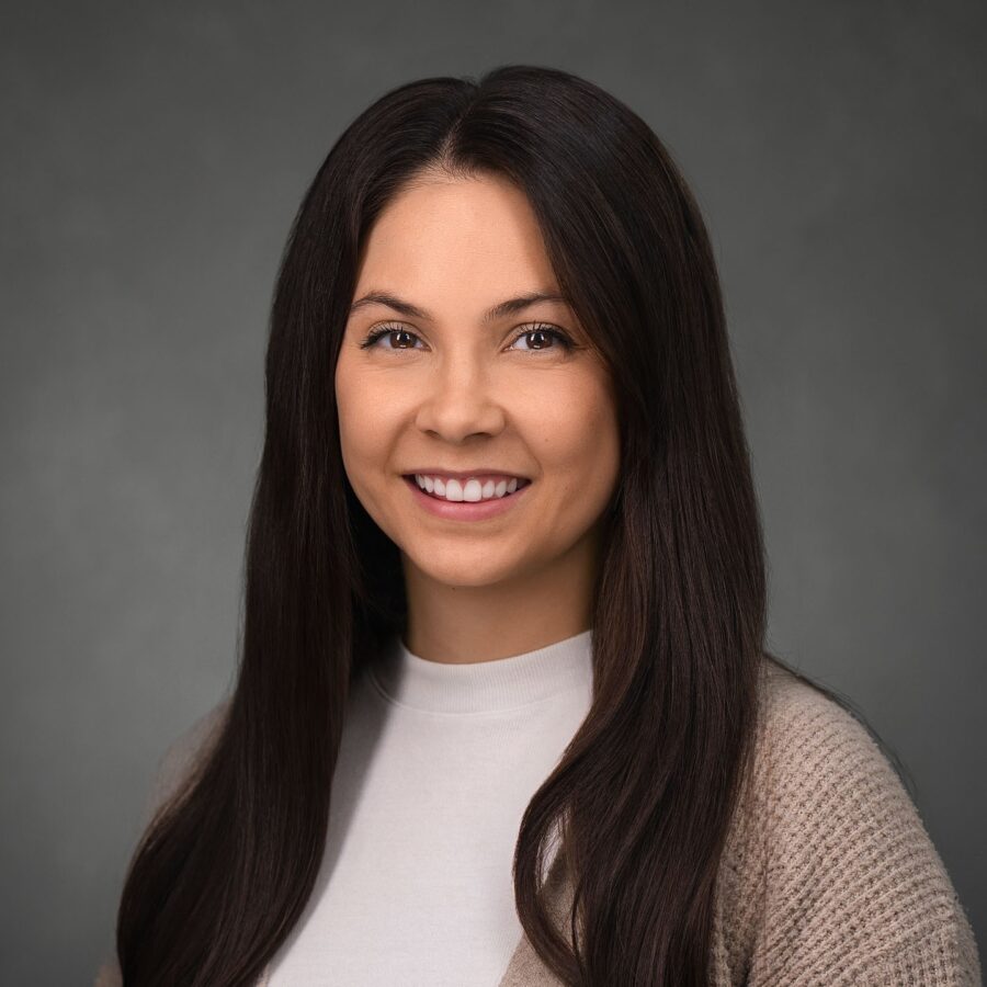 A woman with long dark hair, wearing a light sweater and white top, smiles at the camera against a plain gray background.