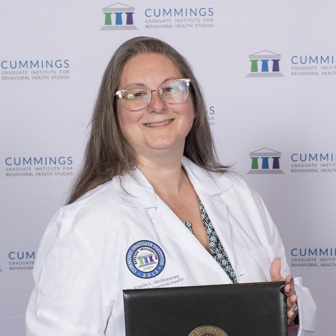 A woman with long brown hair and glasses smiles at the camera. She wears a white lab coat with a badge and holds a black diploma cover. The backdrop displays the Cummings Graduate Institute logo.