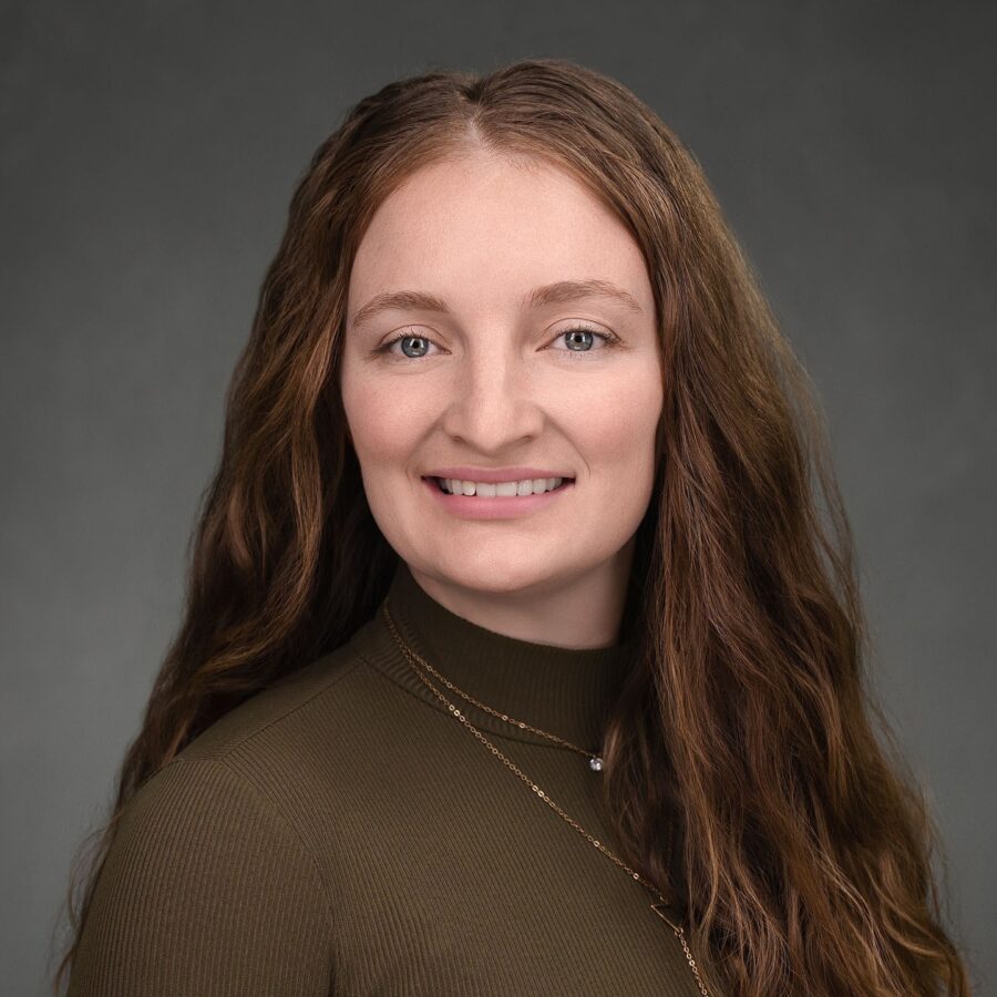 A woman with long wavy brown hair, wearing a dark olive green top and a necklace, poses in front of a gray studio background.
