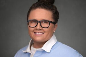 Person with glasses and a topknot bun wearing a light blue collared shirt smiles in front of a plain gray background.