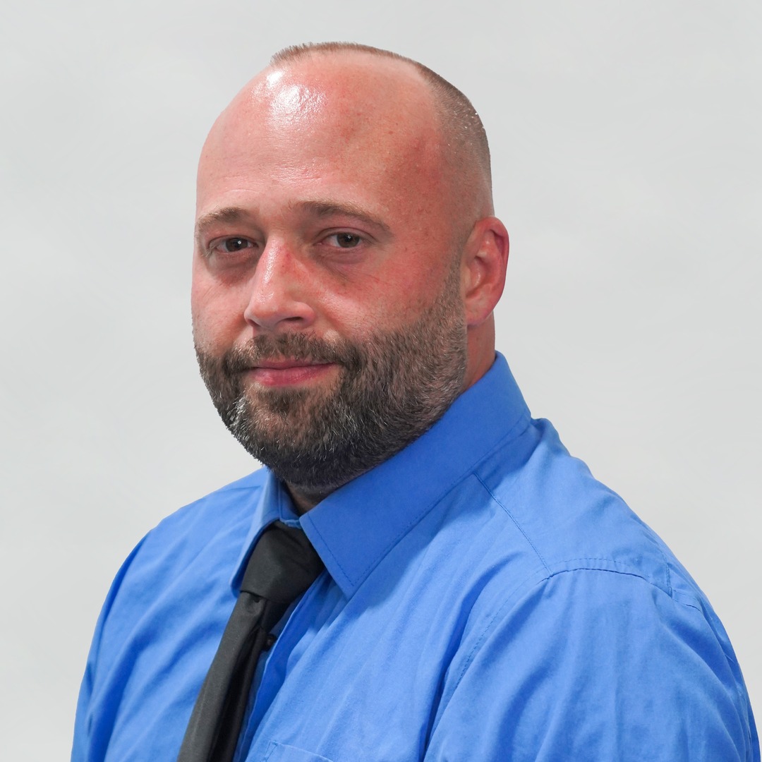 A man with a shaved head and beard, wearing a blue dress shirt and black tie, poses for a portrait against a plain light gray background.