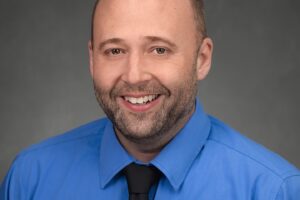 A man with a shaved head and beard, wearing a bright blue dress shirt and black tie, smiles in front of a gray studio background.