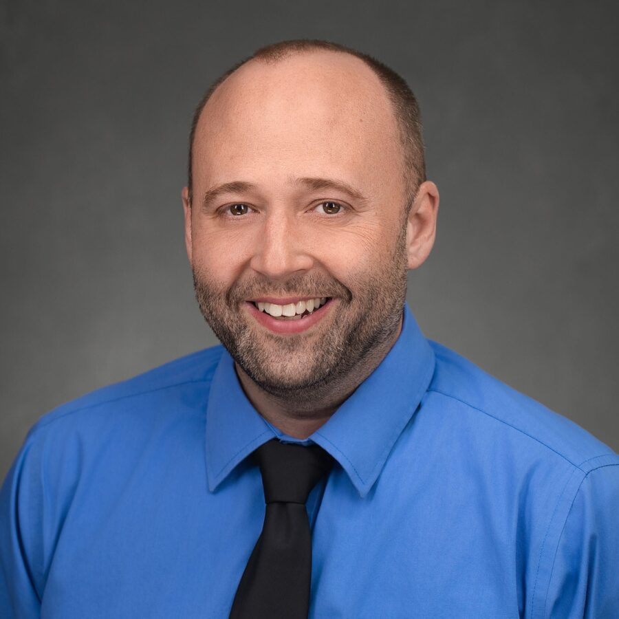 A man with a shaved head and beard, wearing a bright blue dress shirt and black tie, smiles in front of a gray studio background.