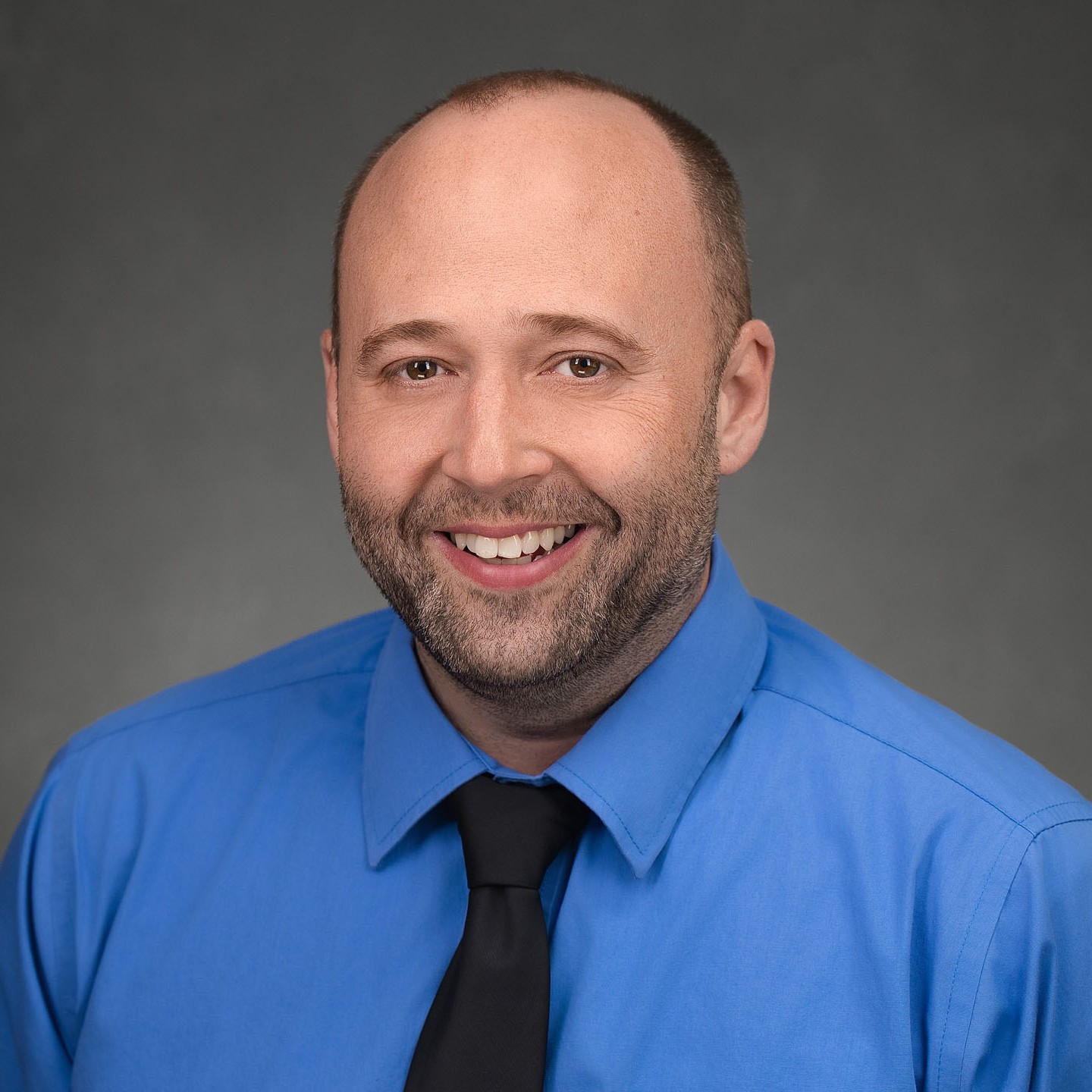 A man with a shaved head and beard, wearing a bright blue dress shirt and black tie, smiles in front of a gray studio background.