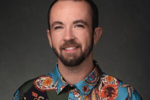 Dr. Tyler Titus with short dark hair and a trimmed beard smiling, wearing a colorful paisley-patterned shirt, against a dark background.