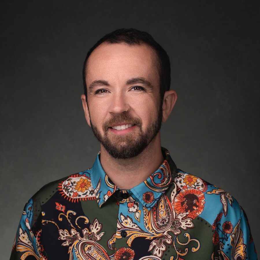 Dr. Tyler Titus with short dark hair and a trimmed beard smiling, wearing a colorful paisley-patterned shirt, against a dark background.