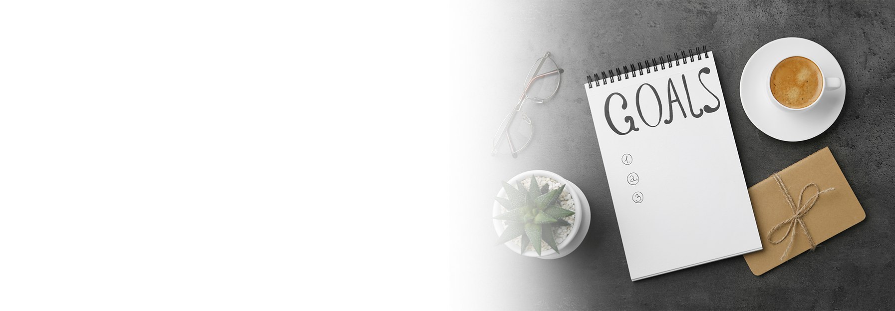 A notepad with GOALS written on it sits on a gray surface beside a cup of coffee, a small potted plant, eyeglasses, and a brown envelope. The left side of the image is white and faded.