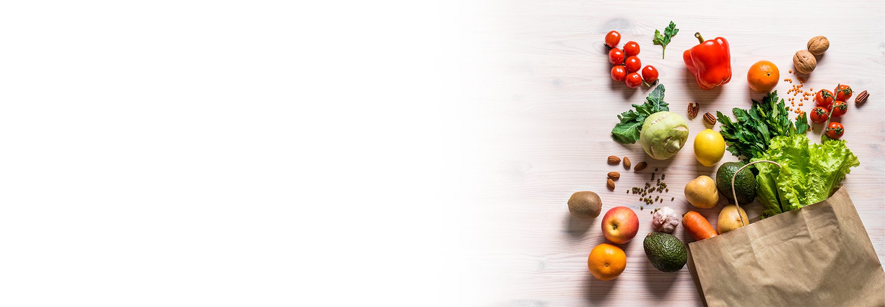 A paper bag filled with fresh vegetables and fruits, including lettuce, avocado, potatoes, tomatoes, apples, orange, walnuts, and a red bell pepper, spilling onto a light wooden surface with empty space on the left.