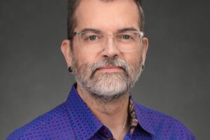 A person with short brown hair, glasses, a beard, and earrings, wearing a blue patterned shirt, poses against a dark gray background.