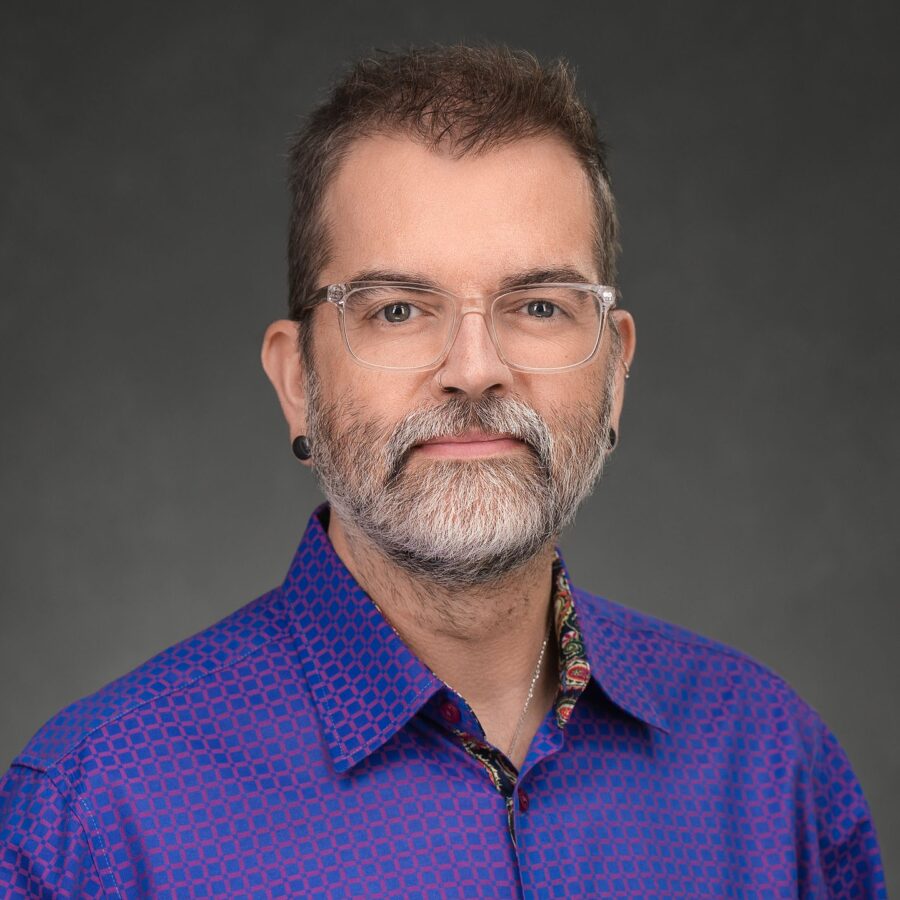 A person with short brown hair, glasses, a beard, and earrings, wearing a blue patterned shirt, poses against a dark gray background.
