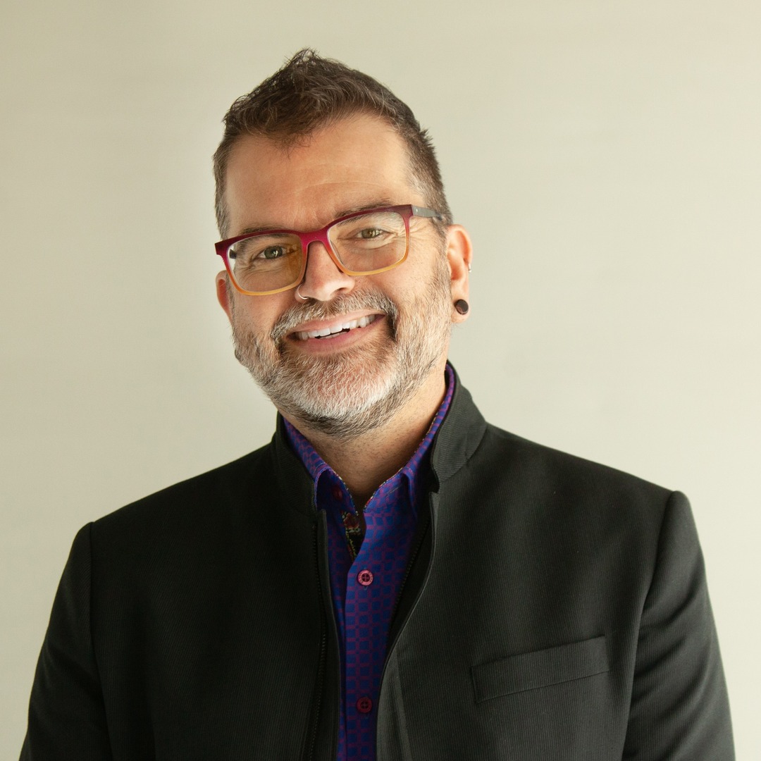 A man with short brown hair, a beard, and glasses with red frames, wearing a black jacket over a purple shirt, smiles in front of a plain light background.