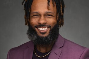 A man with a beard and dreadlocks smiles while wearing a purple blazer, black shirt, and gold chain against a plain dark background.