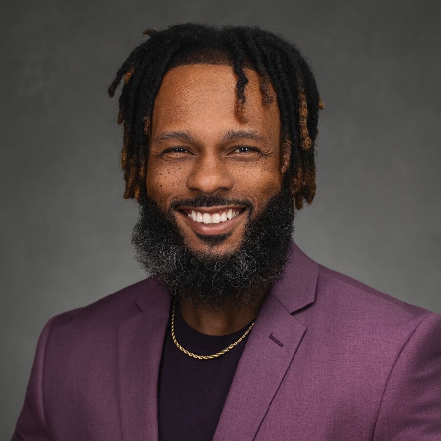 A man with a beard and dreadlocks smiles while wearing a purple blazer, black shirt, and gold chain against a plain dark background.