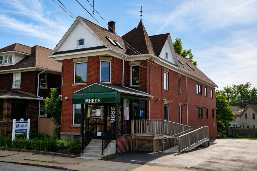 A two-story brick building with a green awning labeled MHEDS, an accessible ramp, and a small sign for MHEDS Health Center on the lawn.
