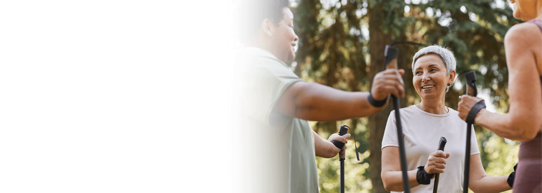 Three people smiling and holding walking poles while exercising outdoors in a wooded area.